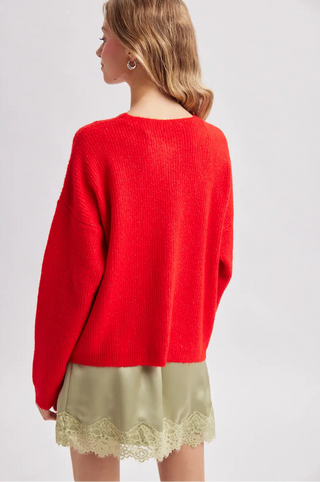 A woman shown from the back wearing a bright red knit sweater and a light olive green satin skirt with a lace hem, against a plain light gray studio background.
