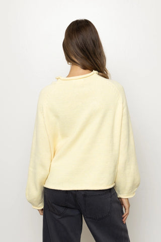 A woman viewed from behind wearing a pale yellow long-sleeve knit top and dark gray pants against a plain white background.