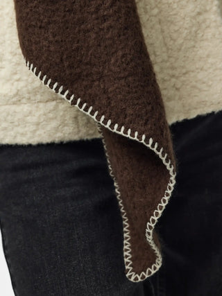 Close-up of a brown textured garment or fabric draped over a cream sherpa-like layer, with dark denim visible below and a white zigzag stitched edge.