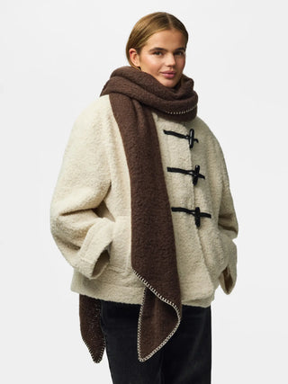 A model wearing a cream-colored bouclé duffle coat with black toggle closures, a dark brown knit scarf, and dark pants, photographed against a light gray studio background.