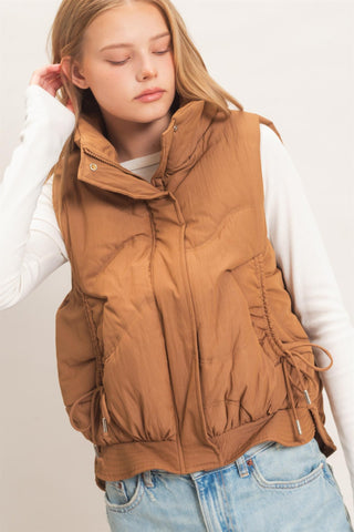 A young woman wearing a brown puffer vest over a white long-sleeve shirt and light blue jeans, photographed against a plain light background.