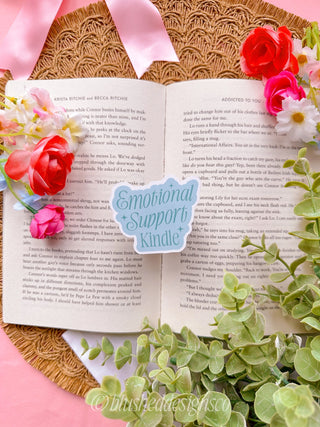Open book with 'Emotional Support Kindle' sticker surrounded by flowers on a pink background