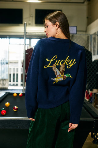 Person wearing a navy blue sweatshirt with 'Lucky' and a duck design, standing in front of a pool table.