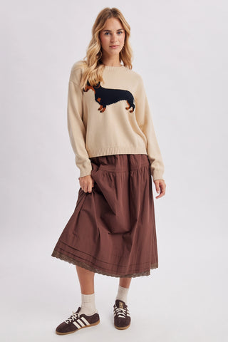 A woman modeling a cream-colored knit sweater with a black and brown dachshund graphic, paired with a brown midi skirt, white socks, and brown sneakers, against a plain light gray background.