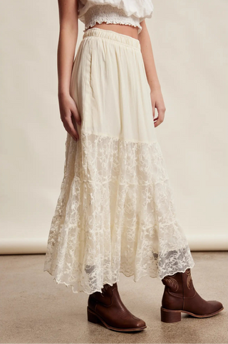 An off-white tiered maxi skirt with a lace hem, worn by a model with brown leather ankle boots against a neutral beige background.