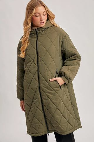 A woman wearing a long olive green quilted puffer coat with a diamond-stitched pattern, black front zipper, and side pockets, photographed against a light gray studio background.