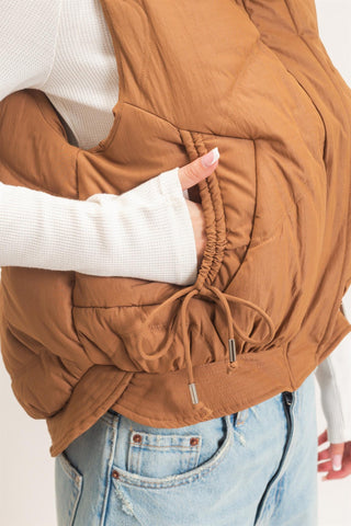 A close-up of a person wearing a brown puffer vest over a white ribbed long-sleeve top and light blue jeans, with the hand reaching into a side pocket.