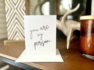 Card with 'you are my person' text on a wooden surface with a candle and decorative items.