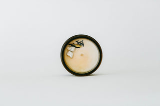 A small round tin candle with a dark rim and pale cream wax, featuring a few decorative stones and a black accent on the lid, set against a plain white background.