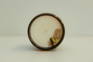 A top-down view of a round jar candle with a dark rim, filled with creamy off-white wax and featuring a small embedded botanical element near the bottom right.