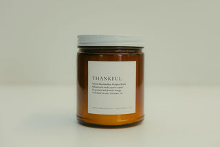 An amber glass jar candle with a white lid and a white label reading 'THANKFUL' on a light gray background.
