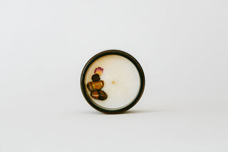 A round glass jar candle with a dark rim, filled with white wax and decorated with small amber beads and a dried pink flower, set against a plain white background.