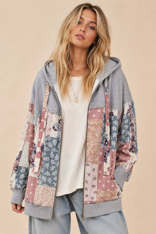 A woman wearing a gray zip-up hoodie with a colorful patchwork pattern of floral and paisley fabric panels, layered over a white t-shirt and light-wash jeans, set against a solid beige background.