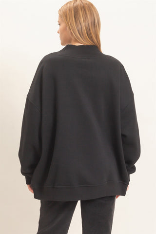 A woman viewed from the back wearing an oversized black sweatshirt and dark pants against a plain light background.
