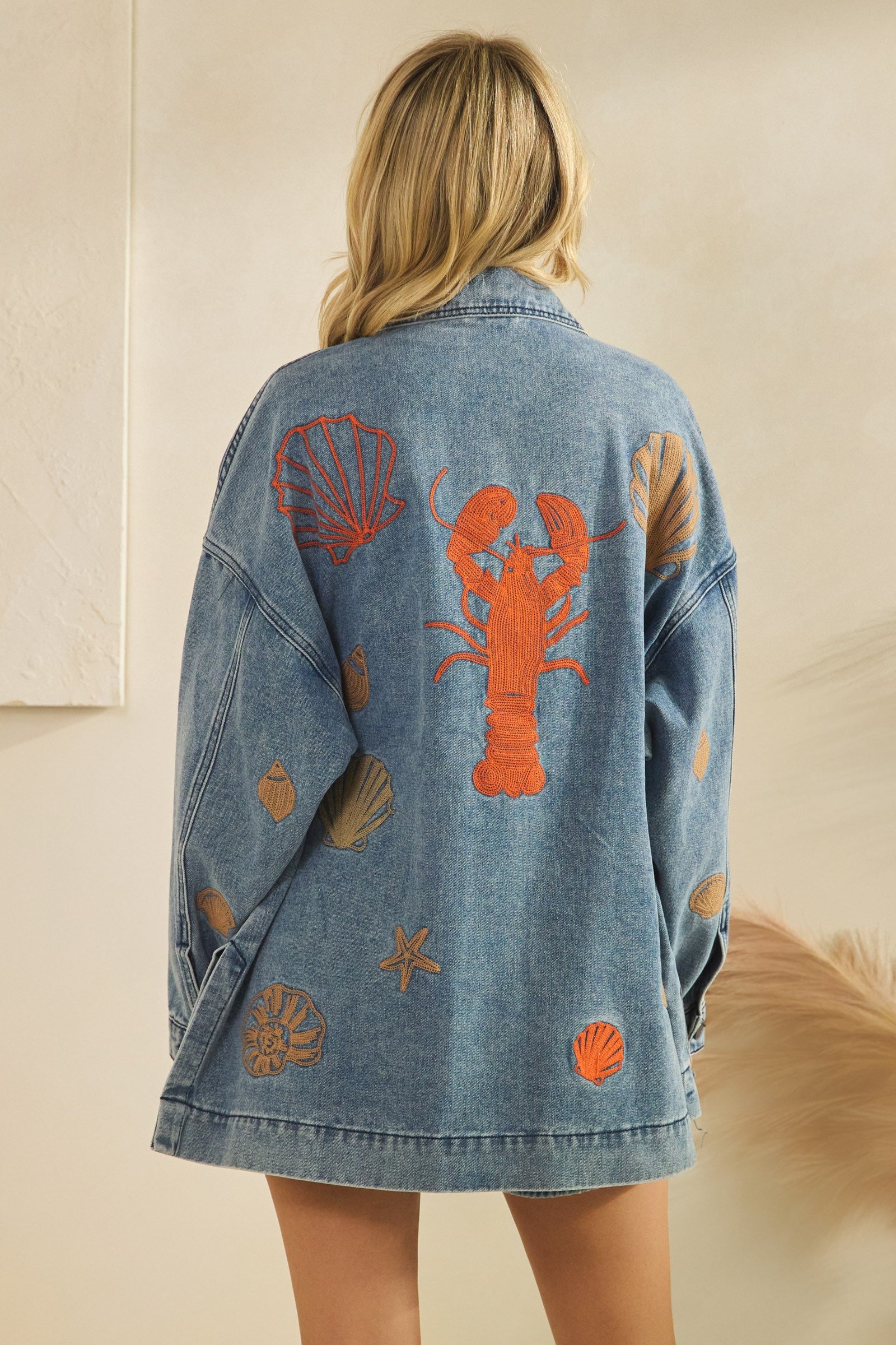 Denim jacket with lobster and shell designs on a neutral background