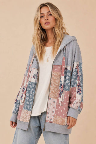 A woman wearing a gray zip-up hoodie with a colorful patchwork floral and geometric print on the front and sleeves, styled over a white t-shirt and light blue jeans against a solid beige background.