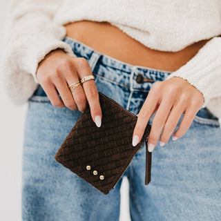 A close-up of a person holding a dark brown woven zip-around wallet with gold-tone studs, wearing a white fuzzy cropped sweater and blue jeans.