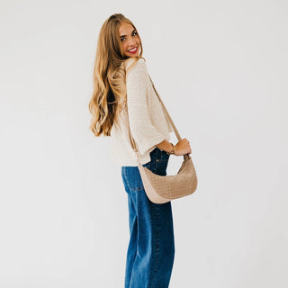 A woman with long blonde hair is shown from behind wearing a cream knit sweater, blue jeans, and carrying a beige woven crescent shoulder bag against a plain white background.