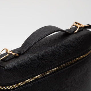 Close-up of a black pebbled leather bag with a top handle, gold-tone hardware, and a gold zipper on a light gray background.
