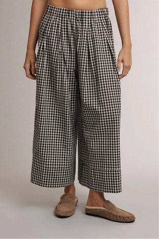 A person wearing loose black-and-white gingham cropped pants with side pockets and tan slip-on mule shoes, photographed against a plain light gray studio background.