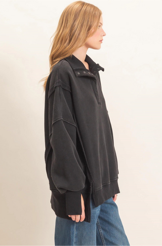 A woman in profile wearing a black oversized quarter-zip sweatshirt and blue jeans, photographed against a plain off-white studio background.