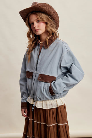 A woman wearing a light blue cropped jacket with brown leather-like trim and pockets, a brown hat, and a brown skirt, photographed against a plain beige studio background.