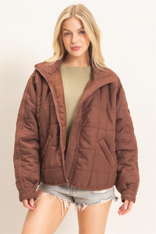 A blonde woman wearing a brown quilted puffer jacket over a beige ribbed sweater and denim shorts, standing against a plain off-white studio background.