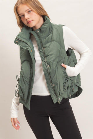 A woman wearing an olive green puffer vest over a white long-sleeve top and black pants, photographed against a plain light background.