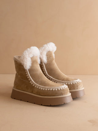 A pair of beige suede ankle boots with white faux-fur trim and a chunky brown platform sole, set against a warm beige background.