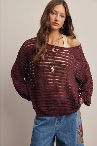 A woman wearing a loose, off-the-shoulder knit sweater in a deep burgundy or maroon color, paired with blue jeans and layered gold necklaces, against a plain light gray studio background.