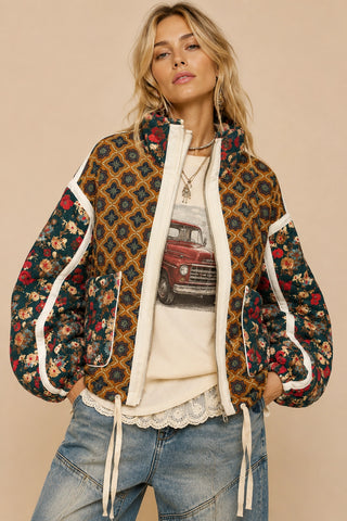 A woman models a bohemian-style patchwork quilted jacket with a geometric pattern on one side and a red floral print on the other, featuring cream-colored piping and a cream zipper. She is wearing a vintage truck graphic t-shirt and blue jeans against a solid beige background.