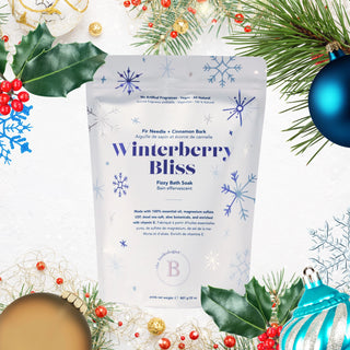 A stand-up pouch of Winterberry Bliss Fizzy Bath Soak centered on a white background, surrounded by holiday decorations including pine branches, holly leaves, red berries, gold ornaments, and blue baubles.
