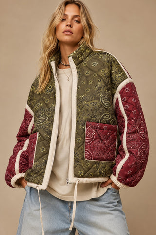 A woman wearing a quilted patchwork jacket with an olive green paisley pattern on the body and sleeves, burgundy paisley sleeves, and cream ribbed trim, styled with a cream t-shirt and light blue jeans against a beige background.