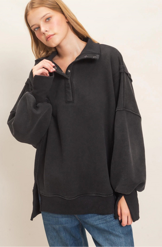 A woman wearing an oversized black half-placket sweatshirt with balloon sleeves and side slits, paired with blue jeans, photographed against a plain light beige studio background.