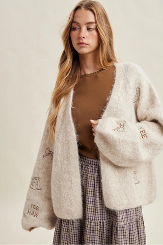 Woman wearing a beige cardigan with embroidered text, brown top, and plaid skirt on a plain background