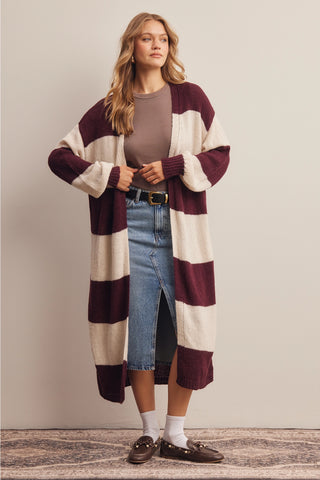 A woman models a long, open-front knit cardigan with wide horizontal stripes in deep burgundy and cream. She is wearing a taupe top, blue jeans, white socks, and brown loafers, standing against a neutral beige wall.