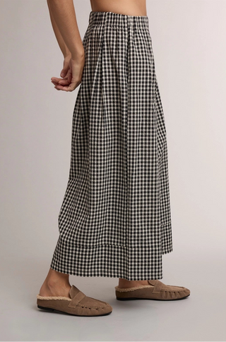 A person wearing loose black-and-white gingham wide-leg pants and tan slip-on mule shoes, shown from the waist down against a plain light gray studio background.