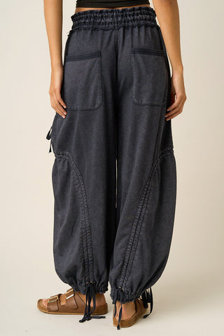 A person wearing dark charcoal gray baggy harem-style pants with an elastic waistband, side pockets, and drawstring ankle cuffs, paired with brown sandals against a plain light beige studio background.