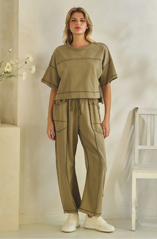 A woman modeling a matching two-piece lounge set in a muted olive green color, featuring a short-sleeve cropped t-shirt and wide-leg drawstring pants, styled with white sneakers in a minimalist indoor setting.