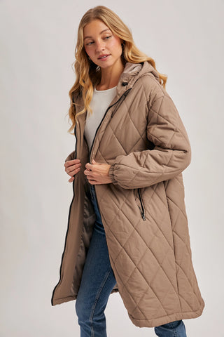 A woman wearing a long quilted hooded puffer coat in taupe brown, styled with a white top and blue jeans, against a plain light gray studio background.