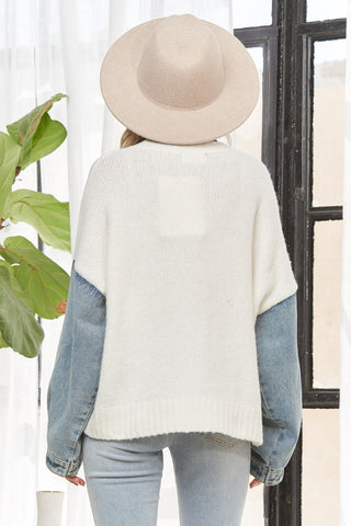Person wearing a white sweater with blue sleeves, light blue jeans, and a beige hat, standing in front of a glass door.
