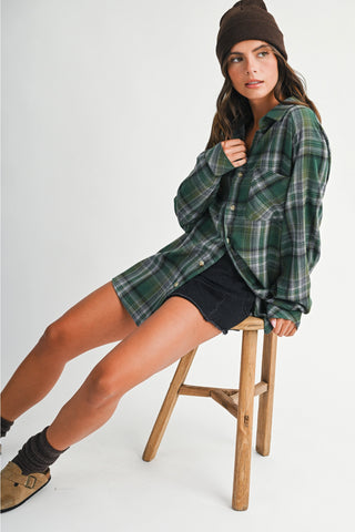 A woman wearing a green and black plaid flannel shirt, black denim shorts, a brown beanie, and brown suede boots sits on a wooden stool against a light gray studio background.