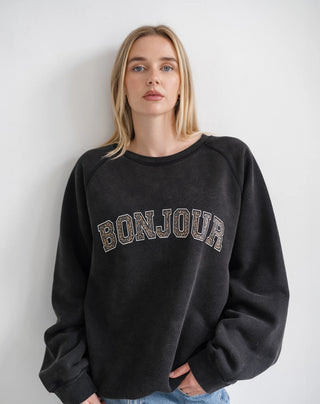 A woman wearing a dark charcoal gray crewneck sweatshirt with 'BONJOUR' written across the chest in a leopard-print pattern, paired with blue jeans against a plain light gray wall.
