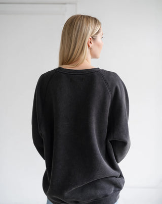 A woman with blonde hair is shown from behind wearing a dark charcoal gray crewneck sweatshirt, photographed against a plain white wall.
