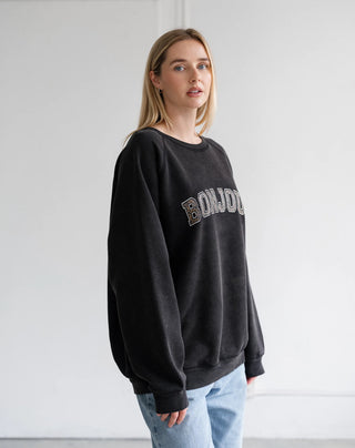 A woman modeling a black crewneck sweatshirt with 'BONJOUR' written across the chest in a metallic gold and silver gradient, paired with light blue jeans against a plain white wall.