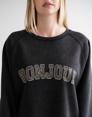 A close-up of a model wearing a black crewneck sweatshirt with 'BONJOUR' arched across the chest in varsity-style block letters filled with a brown and beige leopard print pattern.