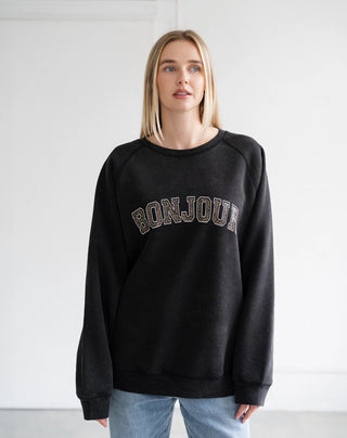 A blonde woman wearing a black crewneck sweatshirt with 'BONJOUR' written across the chest in a textured, metallic gold and silver pattern, paired with light blue jeans, standing against a plain white wall.