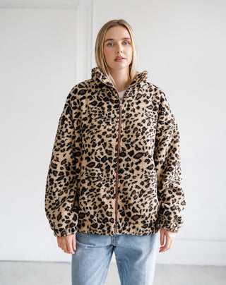 A woman wearing a beige and black leopard print fleece zip-up hoodie jacket with light blue jeans, standing against a plain white wall.