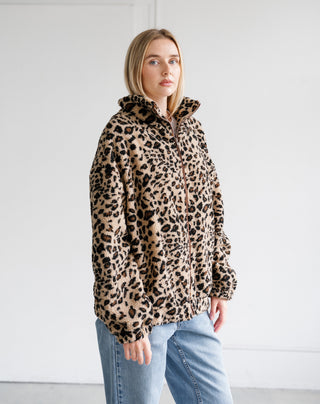 A woman wearing a leopard print fleece jacket with a high collar and front zipper, paired with light blue jeans, standing against a plain white wall.