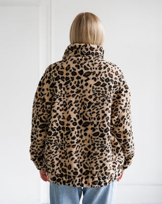 A person viewed from behind wearing a beige and black leopard-print fleece jacket with light blue jeans, standing against a plain white wall.